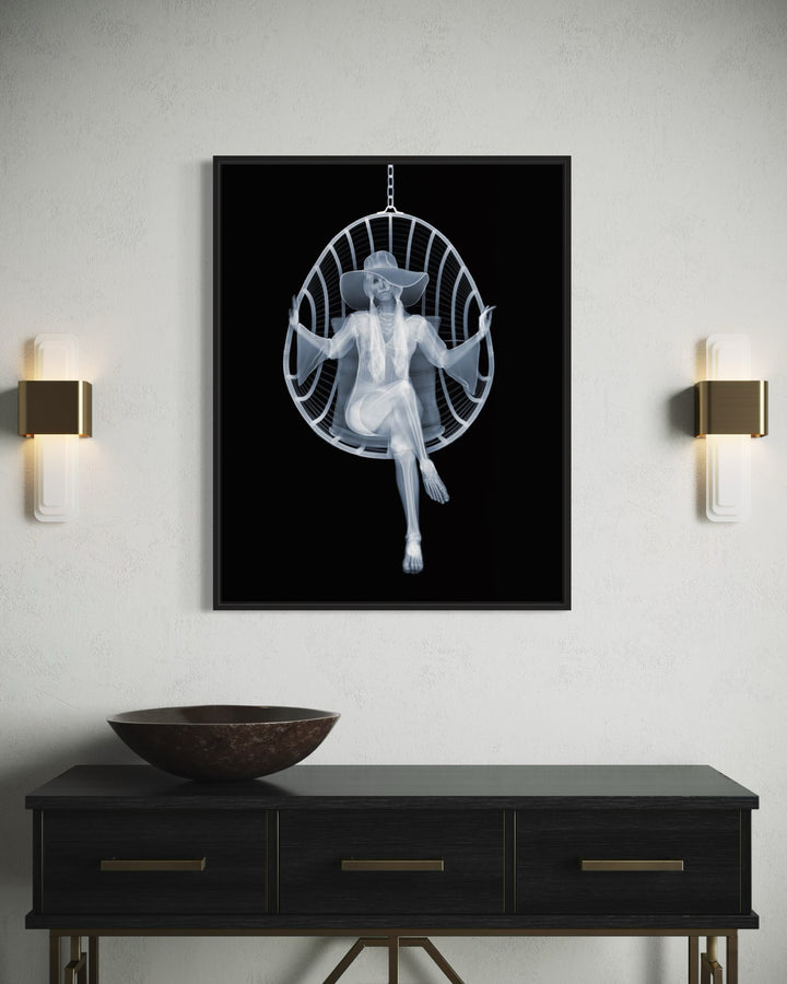 Framed Chromaluxe black and white x-ray photograph by Nick Veasey of a skeleton woman sitting in a swing chair with long pigtails, large hat and boho style dress crossing her legs