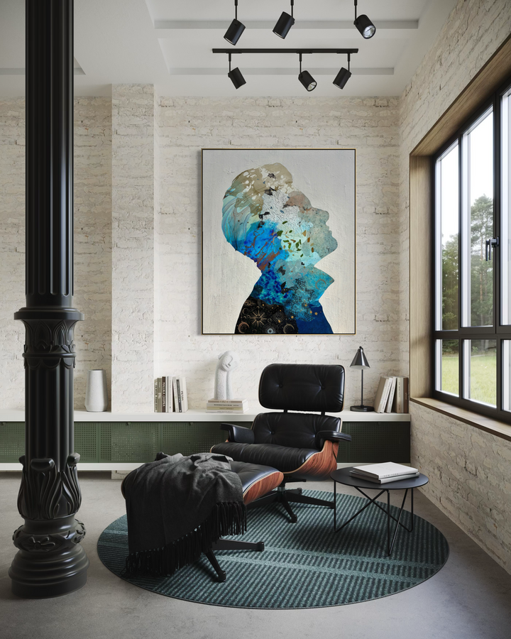 Christopher Peter, Born to Be Blue, acrylic, oil, handmade papers and gold leaf on canvas, 48 x 40 inches. The framed painting of a blue collaged profile silhouette hangs on a whitewashed brick wall above a low shelf with books and a white sculpture, beside a black leather lounge chair and ottoman on a round green rug, with black track lights overhead and a tall window to the right.