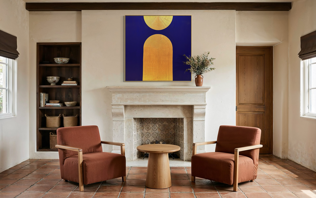 Lee in Cerulean, Halo Vaste II, joint compound and acrylic paint on canvas, 48 x 48 inches. Square deep blue painting with a gold semicircle and gold arch, hung above a stone fireplace in a living room with two rust-colored armchairs, a round wood side table, and built-in shelving.
