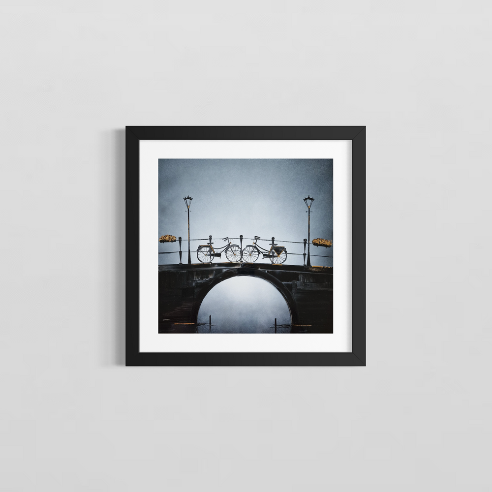 Two bikes facing each other are parked on top of a beautiful bridge over a river with light posts and flower baskets. Painted with black and white Chinese lacquer and gold leaf by Julien Rey. The gold highlights the light in the piece. Framed in a white mat and black frame on a gray wall.
