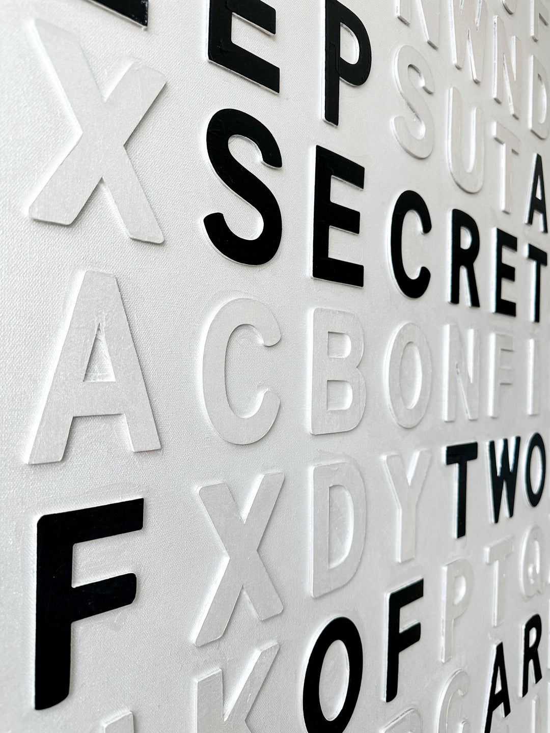 Contemporary 3D text artwork by Chicago artist Steffanie Gerkin titled Keep A Secret 3D, featuring raised black and white wood letters arranged in a structured alphabet grid on a white canvas. The typographic composition spells “Keep a Secret,” “Three May,” and “Two of Them Are Dead,” creating a bold monochrome mixed-media wall art piece with graphic, conceptual, and pop art qualities.