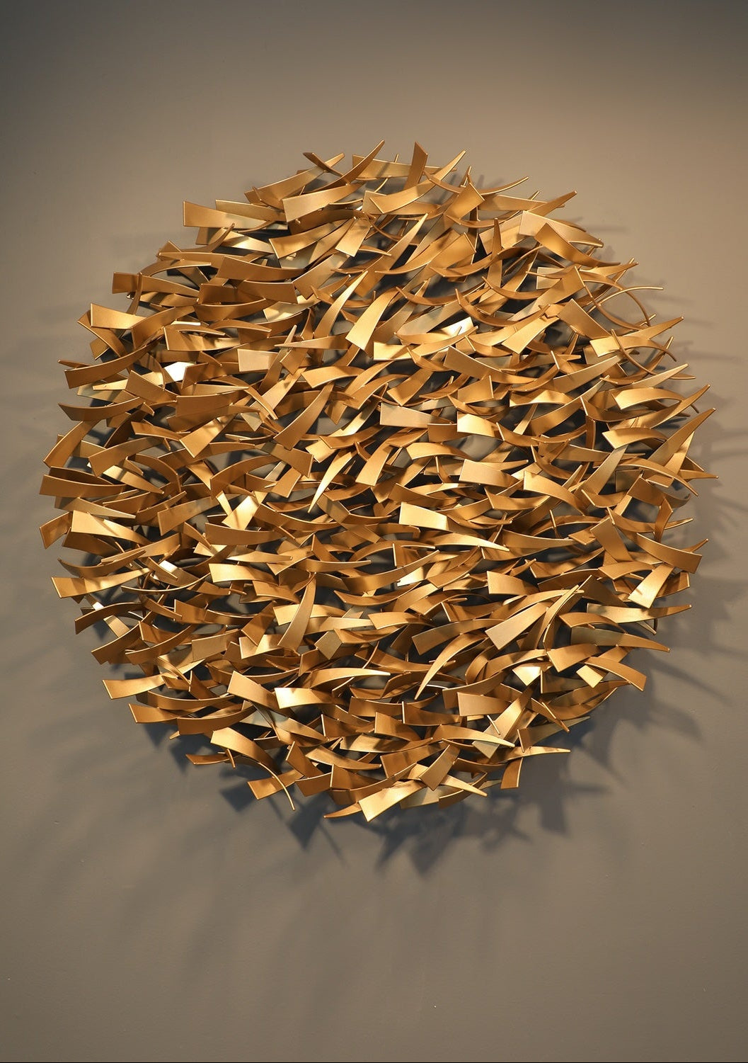 Matt Devine, Avian, powder-coated aluminum, made to order from 11 to 84 inches. Round wall sculpture of many small, curved gold and copper-toned aluminum pieces shaped like bird wings, clustered densely on a beige wall.