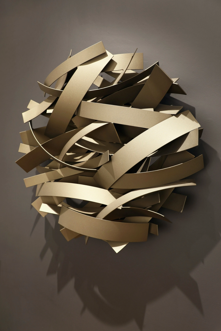 Matt Devine, Mixed Bands, powder-coated aluminum, made to order from 11 to 84 inches. Round wall sculpture of curved champagne aluminum bands in mixed widths, layered in overlapping arcs on a dark taupe wall.