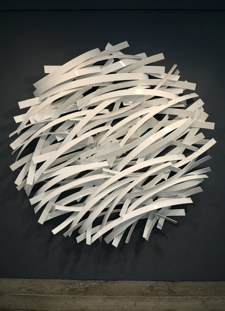 Matt Devine, Thin Bands, powder-coated aluminum, made to order from 11 to 84 inches. Round wall sculpture of thin, curved white aluminum strips layered in overlapping horizontal rows, mounted on a dark grey wall above a concrete floor.