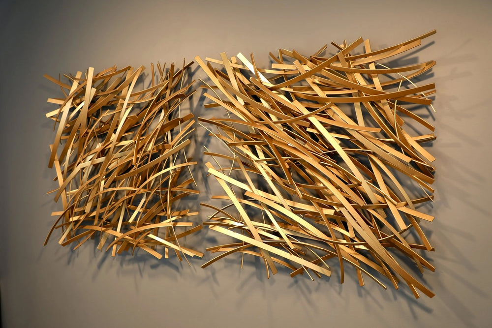 Matt Devine, Thin Bands, powder-coated aluminum, made to order from 11 to 84 inches. Two side-by-side rectangular wall sculptures of thin gold and champagne aluminum strips layered in diagonal crisscross patterns on a beige wall.
