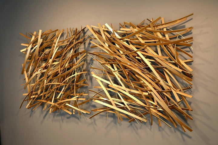 Matt Devine, Thin Bands, powder-coated aluminum, made to order from 11 to 84 inches. Two side-by-side rectangular wall sculptures of thin gold and champagne aluminum strips layered in diagonal crisscross patterns on a beige wall.