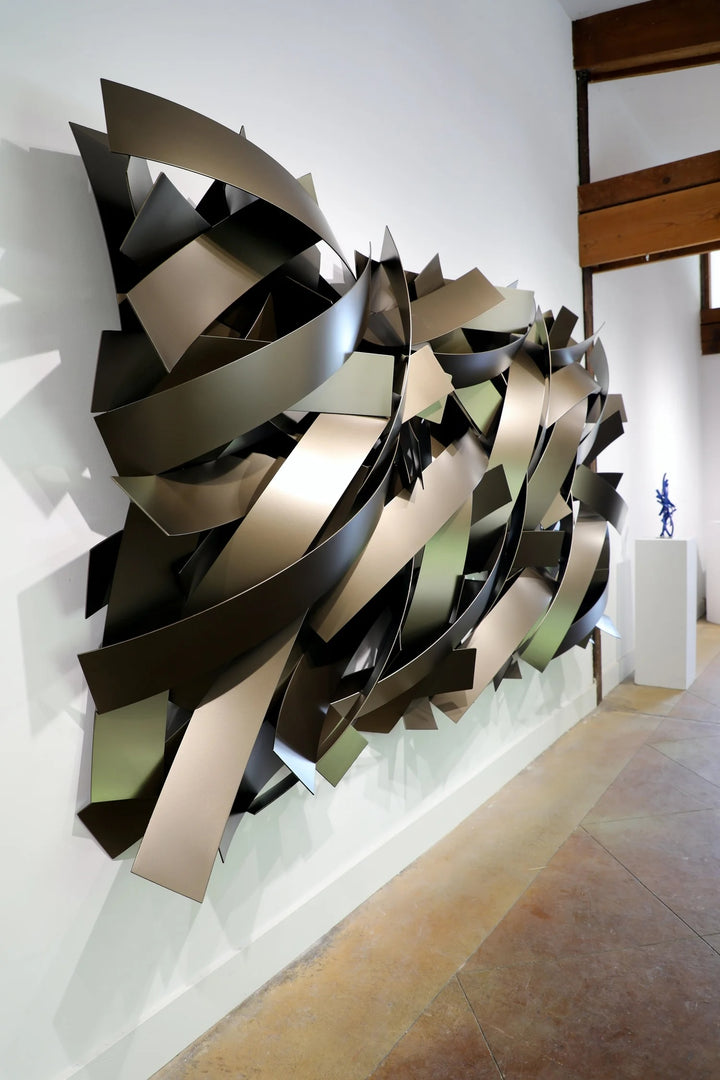 Matt Devine, Wide Bands, powder-coated aluminum, made to order from 11 to 84 inches. Large horizontal wall sculpture of wide, curved champagne and bronze aluminum bands layered in sweeping overlapping arcs, installed on a white gallery wall with a small blue sculpture on a pedestal in the background.