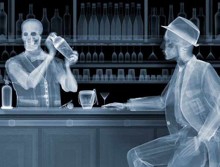 Framed Chromaluxe black and white x-ray photograph by Nick Veasey of two skeletons. A bar tender making a cocktail for a customer sitting at a bar with glasses and bottles behind them