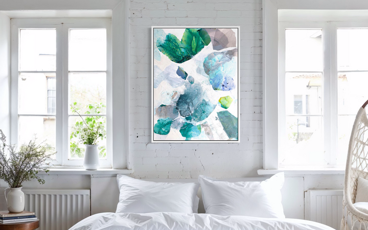 Liz Barber, Sun Bloom 5, acrylic paint, ink, watercolor, and graphite on canvas, 36 x 48 inches. Vertical abstract composition of overlapping rounded shapes in emerald green, teal, sky blue, periwinkle, mauve gray, and lime green scattered across a white background, shown in a white frame on a white brick wall between two windows above a bed with white pillows.