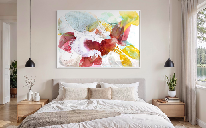 Liz Barber, Tide Pool 2, acrylic paint, ink, watercolor, and graphite on canvas, 72 x 50 inches. Horizontal abstract composition of overlapping translucent shapes in crimson, rose pink, golden yellow, pale blue, and white with fine graphite lines around a white center, shown in a white frame above a bed with a beige upholstered headboard between two black pendant lights.